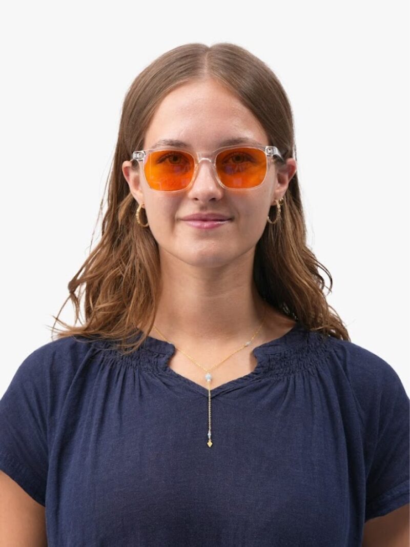 Ung kvinde med gennemsigtige stel og orange solbriller, mørkeblå bluse, guld hoop-øreringe og hengende halskæde