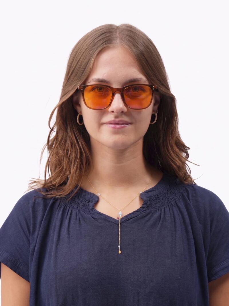 Kvinde med orangetonede solbriller, gyldne øreringe og delikat guldkæde iført navy bluse