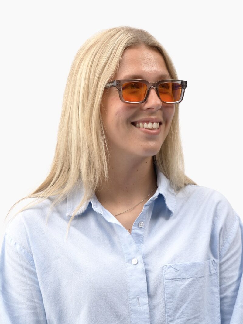 Smilende kvinde med langt lyst hår iført Eclipse 90 gråt transparent stel og orange linser, lyseblå skjorte