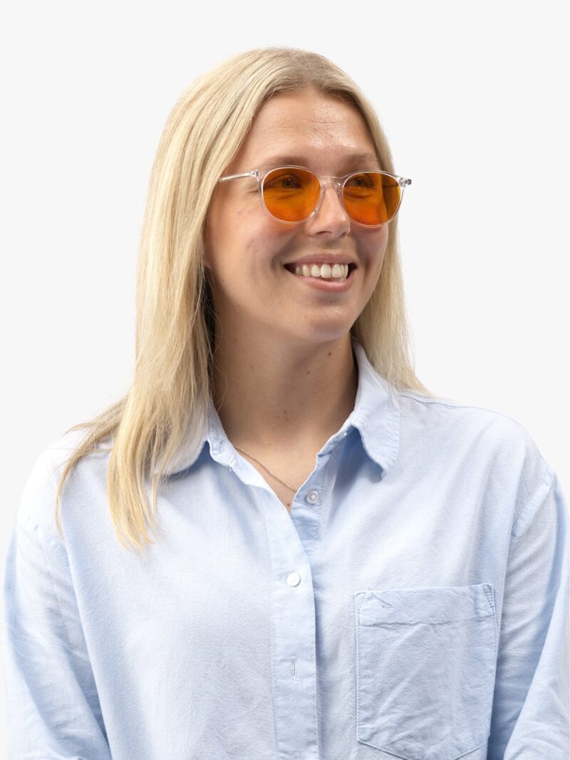Smilende kvinde med langt blondt hår i gennemsigtigt stel med orange linser og lyseblå skjorte
