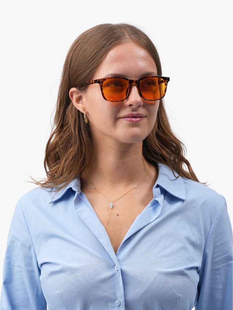 Kvinde iført Obsidian 90 brune solbriller med orange linser, lys blå skjorte og guldsmykker