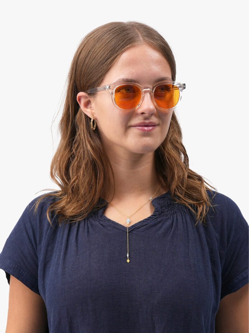 Ung kvinde med klare stel-solbriller med orangetonede linser, navy T-shirt, gyldent vedhængssmykke og skulderlangt brunt hår