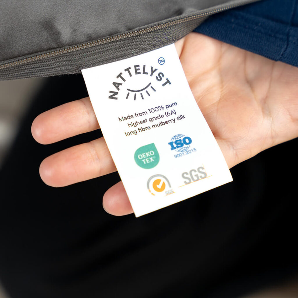 Hånd holder hvidt mærke med NATTELYST-logo, tekst om 100% mulberry-silke og OEKO-TEX, ISO og SGS-certifikater