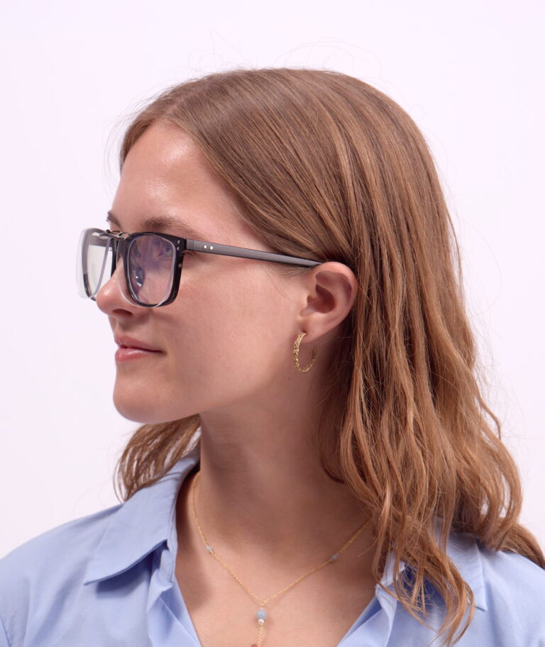 Kvinde i profil med sorte briller, gyldne hoop-øreringe og guldkæde med blåt vedhæng, iført lyseblå skjorte