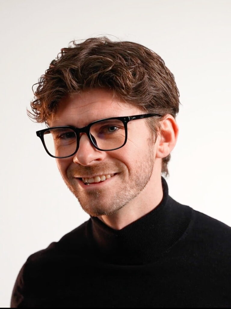 Smilende mand med brunt krøllet hår, sorte briller og sort rullekrave mod lys baggrund