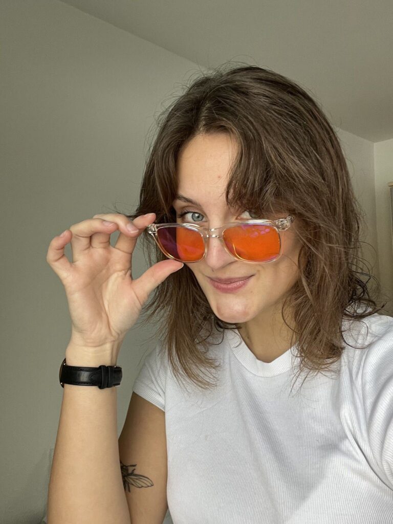 Smilende kvinde i hvid t-shirt løfter orange spejlbriller, synlig insekt-tatovering og sort armbåndsur