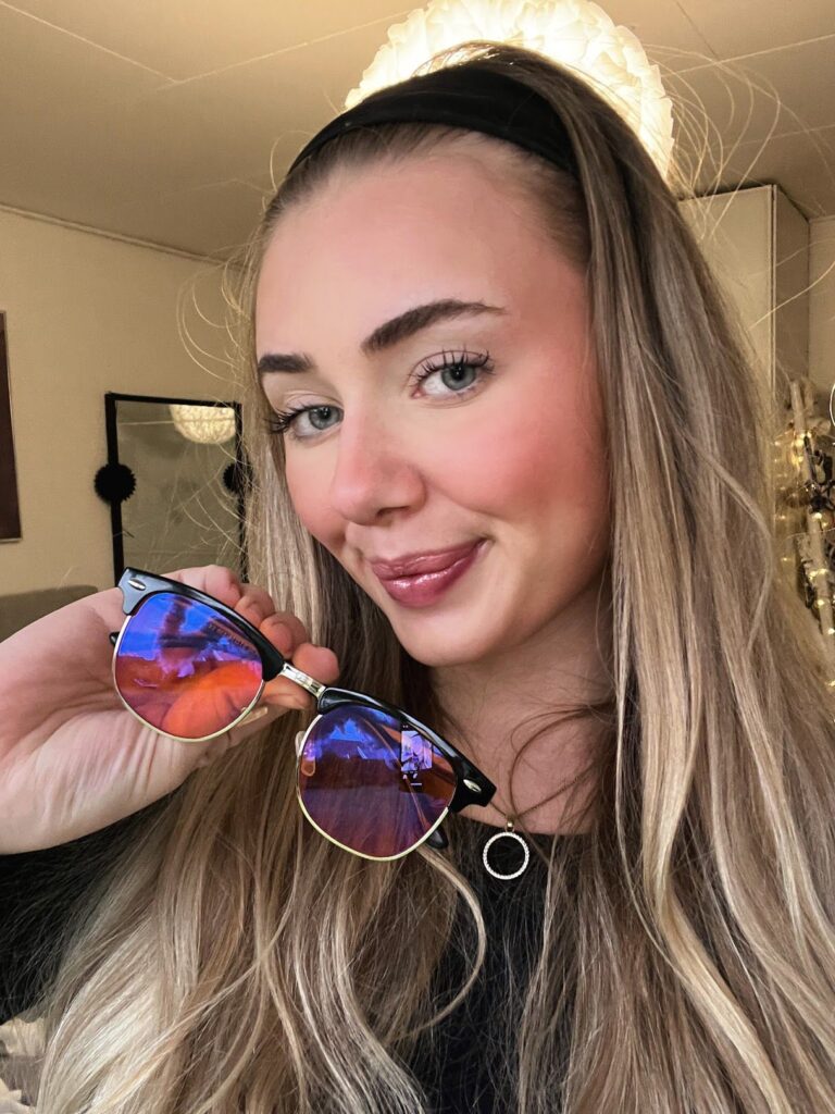 Smilende ung kvinde med langt blondt hår holder blålysbriller med blå-lilla reflekterende linser