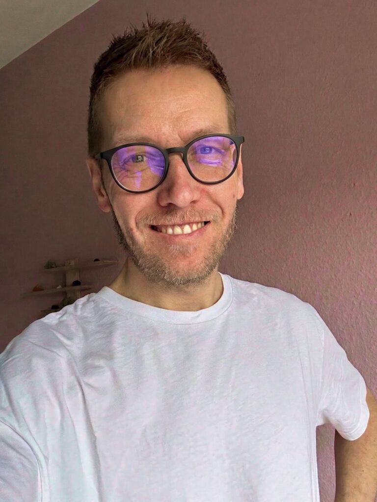 Smilende mand med briller, blå lysreflektion, hvid t-shirt og kort skæg foran lyserød væg